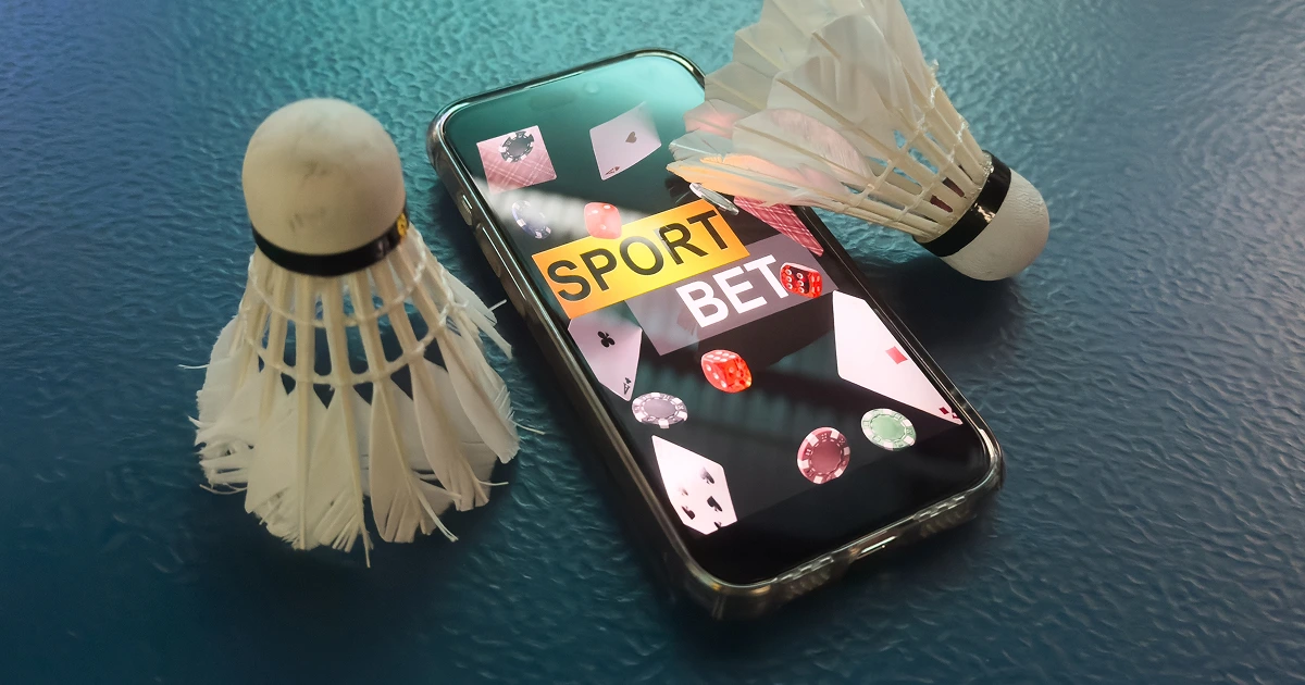 Smartphone with sport bet interface surrounded by badminton shuttlecocks and casino poker chips on a blue surface.
